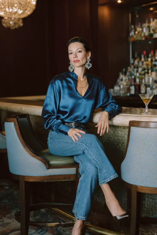 A woman over 40 wears classic blue jeans with a tailored satin blouse tucked in, styled with block-heel pumps and statement earrings