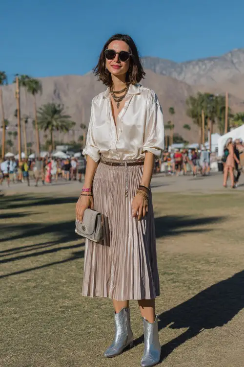 A woman over 40 wears metallic boots with a pleated midi skirt and silky blouse, accessorized with layered chains
