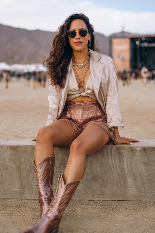 A woman over 40 wears metallic bronze cowboy boots with tailored high-waisted shorts and a silky blouse, finished with a cropped blazer and elegant layered necklaces