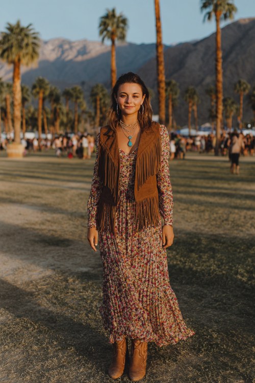 A woman wearing a modest floral maxi dress with long sleeves layered with a brown suede fringe vest, styled with cowboy boots and turquoise jewelry