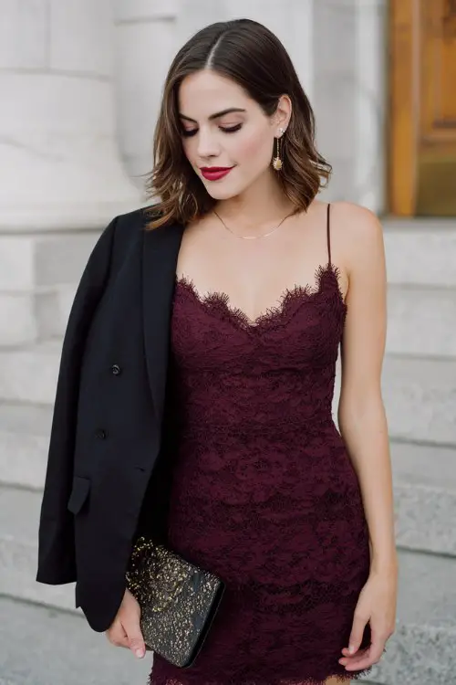 A woman wears a deep burgundy lace cocktail dress with scalloped hem and delicate embroidery, accessorized with a statement clutch and sleek blazer