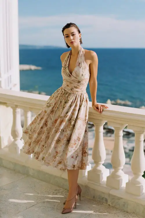 A woman wears a refined spring cocktail dress with pleated skirt, subtle tropical floral pattern and structured bodice, paired with sleek stilettos