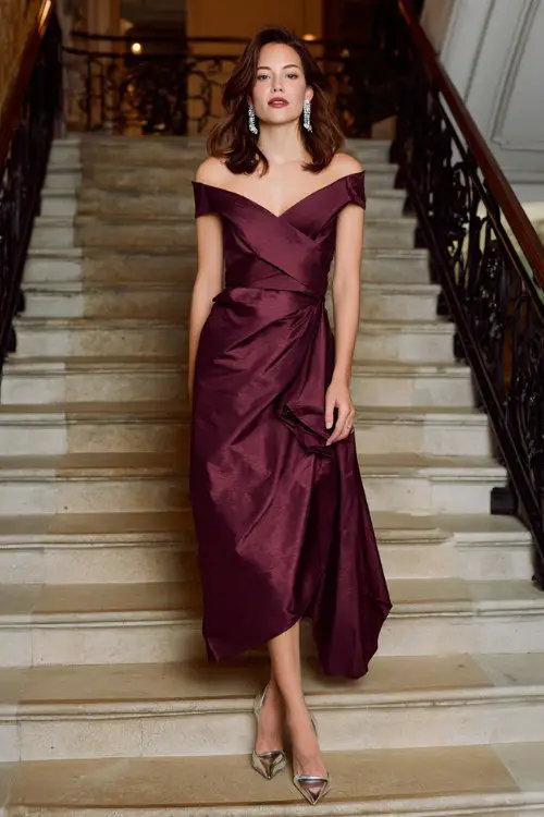 A woman wears a rich plum wrap-effect gown with an asymmetric neckline and satin texture, paired with chrome heels and chandelier earrings