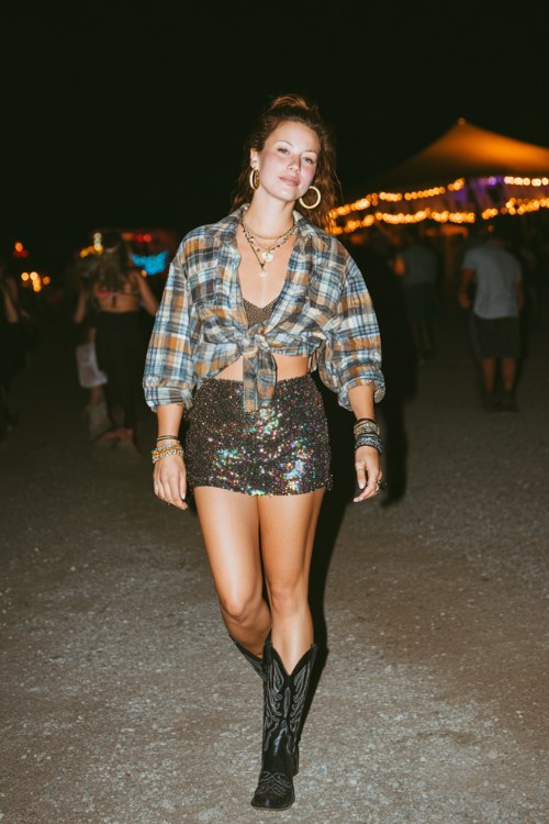 A woman wears black cowboy boots with a shimmering sequin mini skirt and oversized plaid shirt tied at the waist