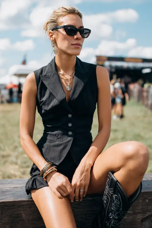 A woman wears black cowboy boots with a tailored vest and matching high-waisted shorts set, styled with bold sunglasses and stacked bangles