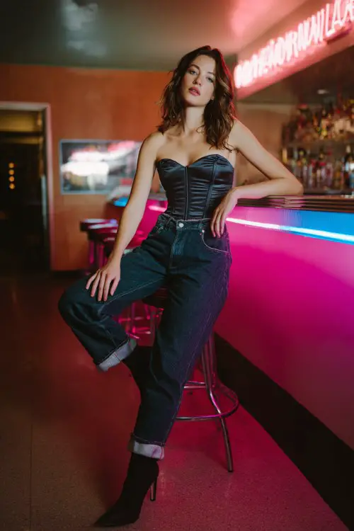 A woman wears dark denim jeans with a corset-style satin top and heeled ankle boots