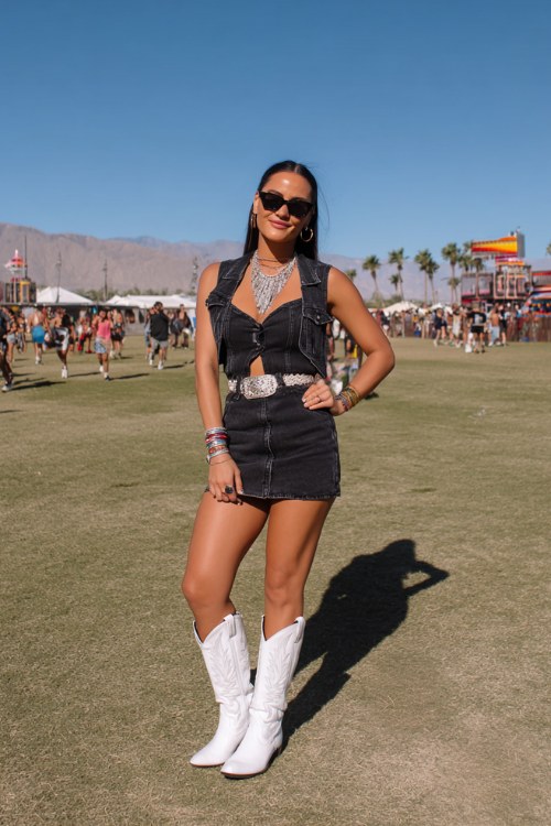 A woman wears glossy white cowboy boots with a cutout bodycon mini dress and cropped denim vest, accessorized with stacked bangles and a bold western belt