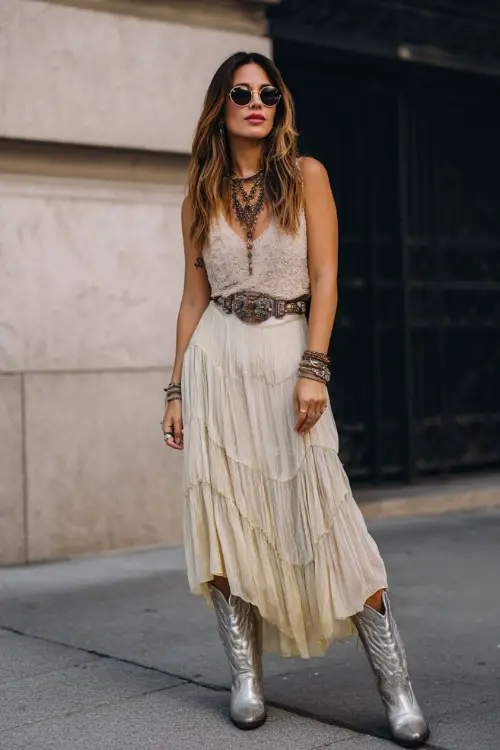 A woman wears metallic cowboy boots with a tiered chiffon maxi skirt and fitted lace top, accessorized with layered necklaces and bold sunglasses