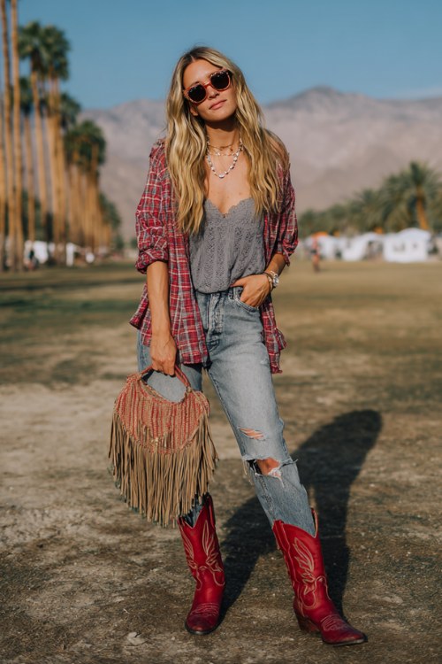 A woman wears red cowboy boots with distressed straight-leg jeans and a lace-trim camisole, layered with a relaxed plaid shirt and a fringe bag