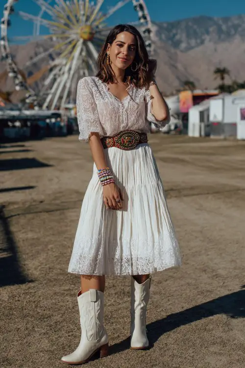 A woman wears white cowboy boots with a soft pleated midi skirt and a lace-trim blouse with long sleeves, styled with a wide western belt and stacked bracelets