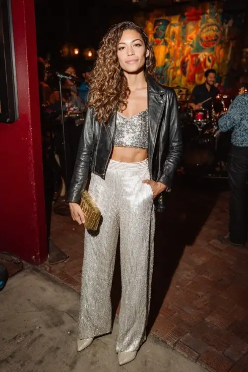 A woman wears wide-leg trousers with a sequined silver top, styled with a cropped leather jacket, pointed-toe boots, and a clutch
