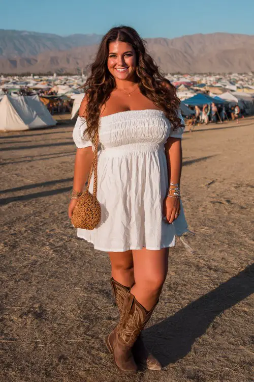 Plus size woman wearing an off-shoulder white boho mini dress, tall cowboy boots, woven crossbody bag, voluminous curls