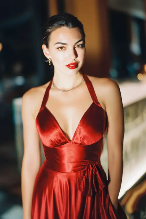 Rocker-inspired full body cocktail guest dress for spring wedding, glossy red satin dress with deep neckline, soft romantic interior background, polished glam makeup