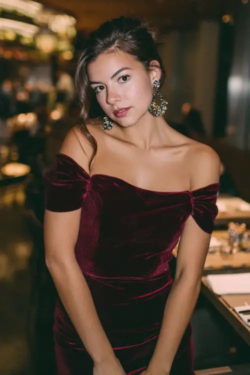 Spring wedding rocker guest outfit, deep burgundy velvet off-shoulder mini dress, statement earrings, warm restaurant lighting, sleek hairstyle, pumps