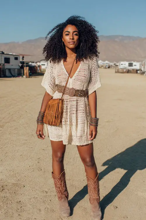 Woman wearing a cream crochet mini dress, brown cowboy boots, fringe bag, stacked bangles