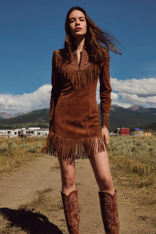 a confident cowgirl woman wearing a fitted brown suede western mini dress with long fringe hem, brown cowboy boots
