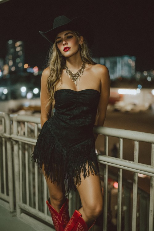 western nightlife fashion look, woman wearing a black fringe mini skirt and black strapless crop top, wide black cowboy hat, statement necklace