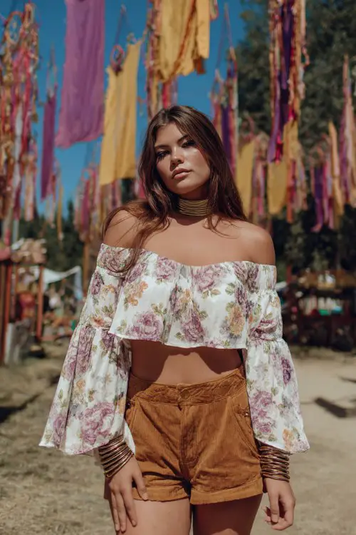 woman in off shoulder ivory floral crop blouse with dusty rose sage green and muted lavender print, loose bell sleeves, high waisted camel tan suede shorts