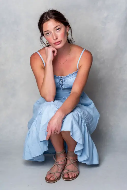 A woman in a light blue smocked sundress with simple jewelry and flat lace-up sandals, styled for a soft casual evening look, simple studio background 