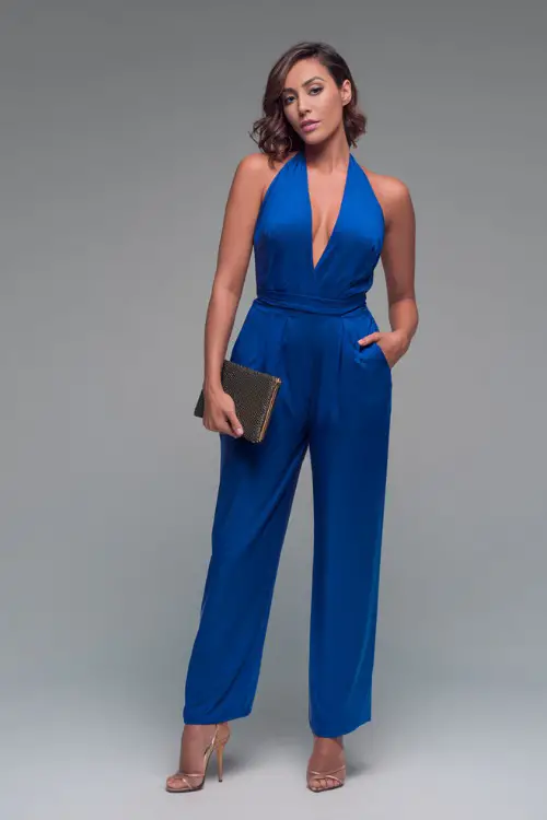 A woman wearing a deep V-neck jumpsuit in bold blue with stiletto heels and a clutch bag, styled for a fashionable summer night out, simple studio background 