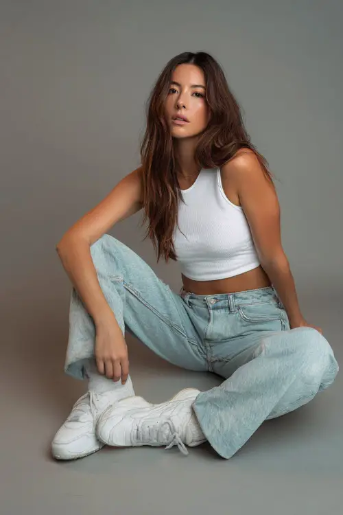 A woman wearing a white cropped tank top with high-waisted light-wash jeans and white sneakers, styled for a casual summer evening, simple studio background 