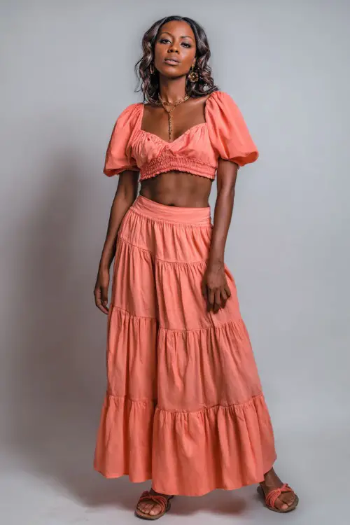 A Black woman wearing a coral crop top with puff sleeves, paired with a flowy tiered maxi skirt and sandals, styled for an elegant summer night look 