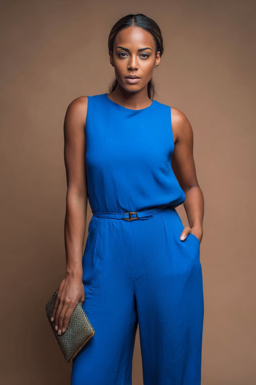 A Black woman wearing a sleeveless jumpsuit in bright cobalt blue with a belt and clutch bag, styled for a sleek and striking summer outfit, simple studio background 