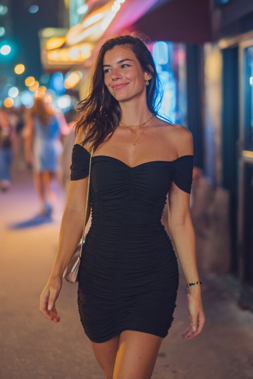A midsize brunette woman wears a casual summer going out outfit with a ruched off-shoulder mini dress with a small shoulder bag and delicate jewelry