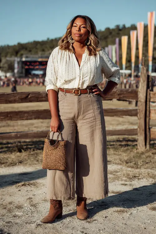 A plus-size woman dressed in linen wide-leg pants, a tucked-in eyelet blouse, and brown ankle cowboy boots, accessorized with statement hoop earrings and a woven bag 