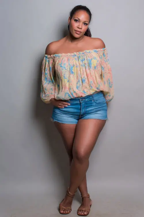 A plus-size woman in a flowy off-shoulder top paired with high-waisted denim shorts and flat sandals, styled for a casual summer night out, simple studio background 
