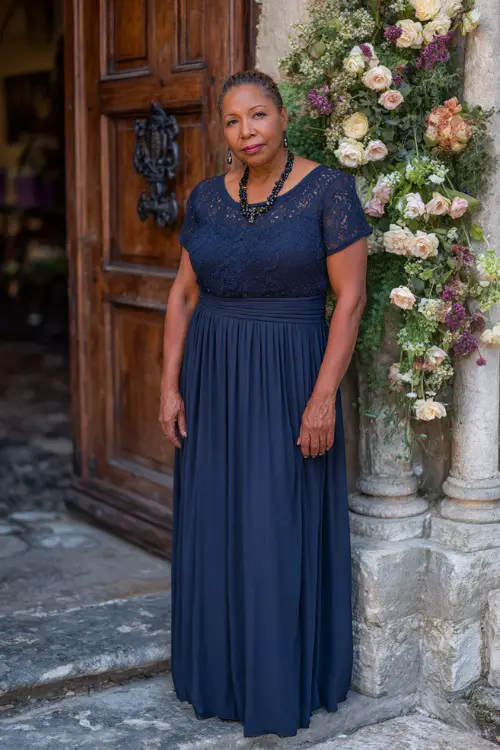 A plus-size woman over 50 wearing a navy blue empire waist maxi dress with short sleeves and a beaded necklace, standing near a classic church entrance with floral decorations 