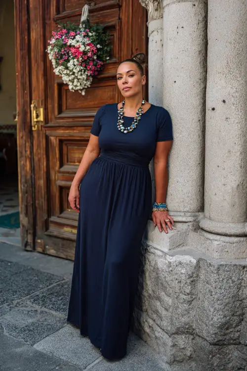 A plus-size woman over 50 wearing a navy blue empire waist maxi dress with short sleeves and a beaded necklace, standing near a classic church entrance with floral decorations 