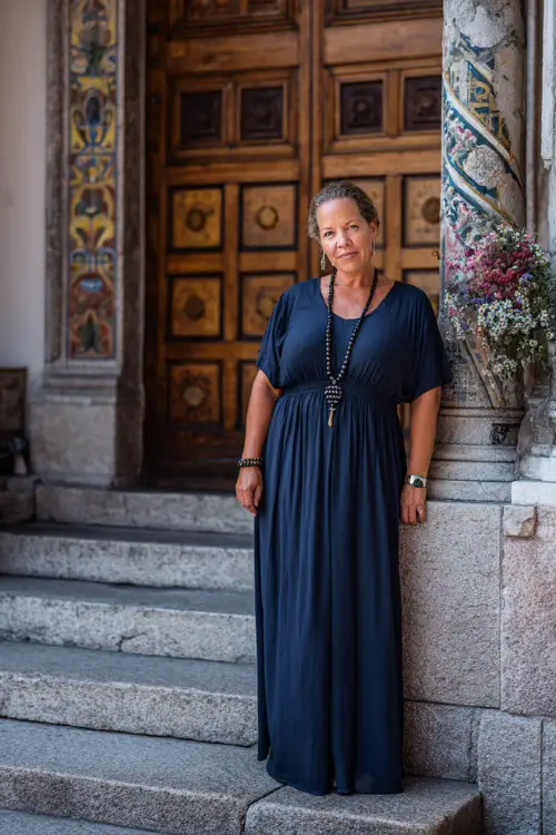 A plus-size woman over 50 wearing a navy blue empire waist maxi dress with short sleeves and a beaded necklace, standing near a classic church entrance with floral decorations
