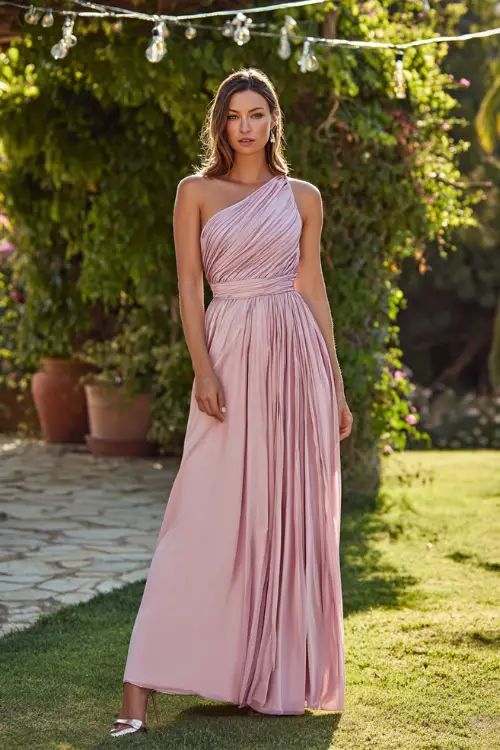 A woman in a blush pink one-shoulder formal gown with pleated details and metallic heels, standing under string lights at a glamorous garden wedding 