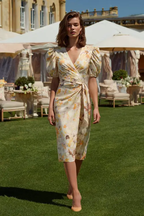 A woman in a floral wrap midi dress with puff sleeves and tan heels, walking across a sunny lawn set up for a modern summer cocktail ceremony
