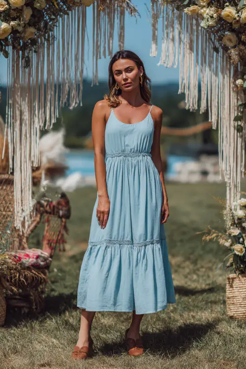 A woman in a light blue sleeveless sundress with a tiered skirt and tan flats, standing near a boho-inspired outdoor wedding arch with macramé details