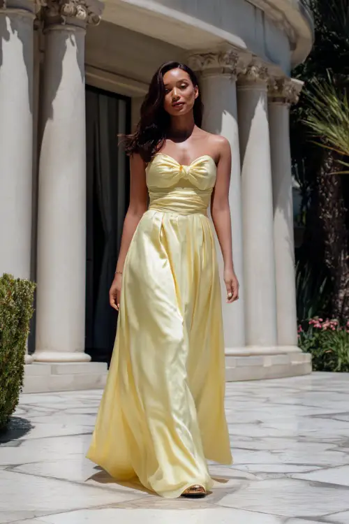 A woman in a pastel yellow satin gown with a sweetheart neckline and minimal accessories, walking across a marble patio at a high-end summer wedding 