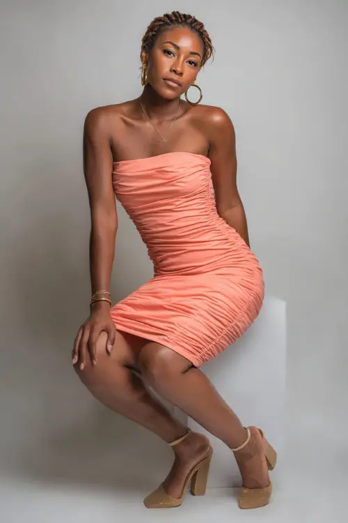 A woman in a strapless ruched bodycon dress in coral with gold hoop earrings and block heels, styled for a fun summer bar outing, simple studio background 