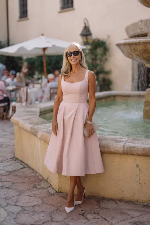 A woman over 50 in a blush pink fit-and-flare midi dress with a square neckline and low block heels, posing by a stone fountain at a summer courtyard wedding 