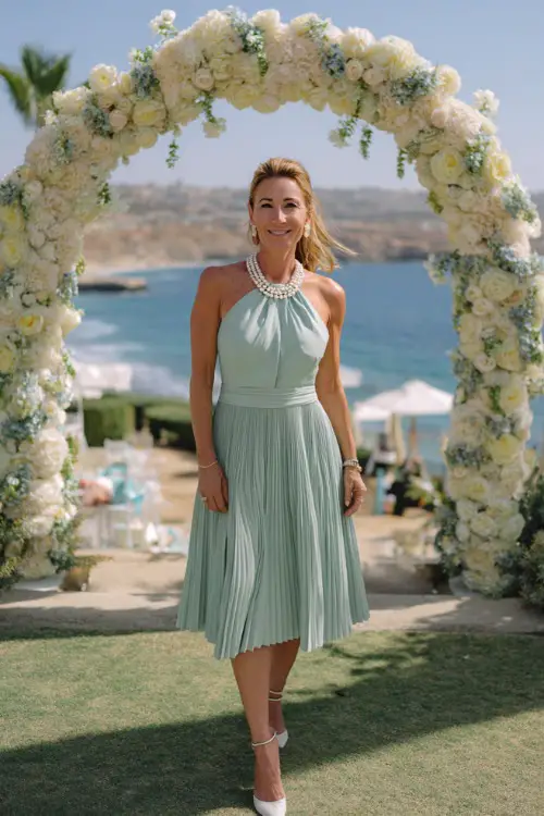 A woman over 50 in a pastel mint green midi dress with pleated skirt and pearl necklace, posing by a decorated entry arch at a coastal cocktail wedding