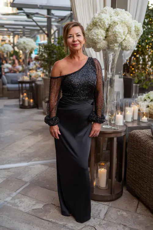 A woman over 50 in a sleek black cocktail dress with sheer sleeves and sequin accents, standing on a patio with white floral arrangements and candlelit décor 
