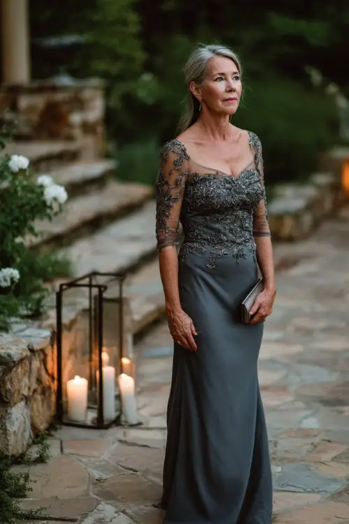 A woman over 50 wearing a charcoal gray embellished gown with illusion sleeves and a subtle mermaid silhouette, posing near a candlelit pathway at a twilight garden wedding 