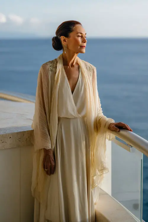 A woman over 50 wearing a flowy off-white chiffon gown with a sheer shawl and low bun hairstyle, standing on a balcony overlooking the ocean