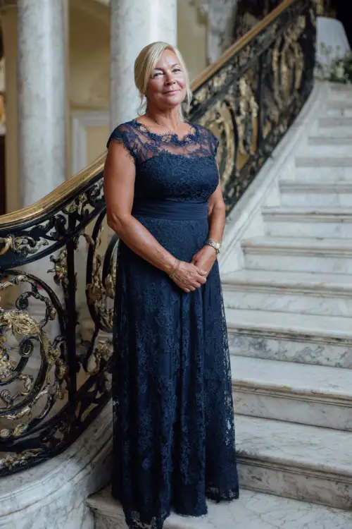 A woman over 50 wearing a navy lace overlay gown with scalloped edges and low heels, standing at the top of a staircase inside a classic estate wedding venue 
