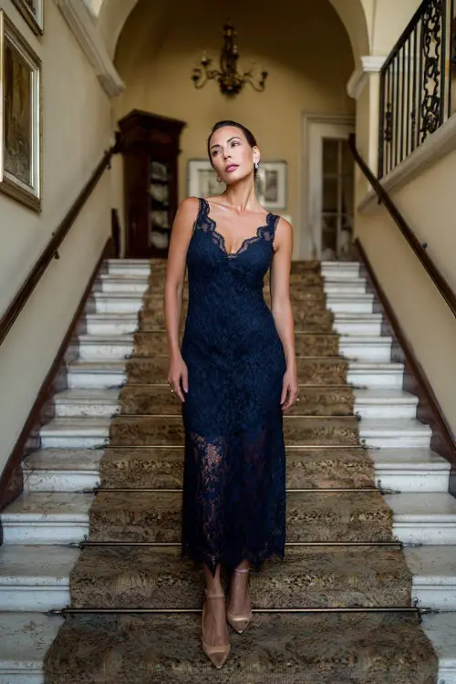 A woman over 50 wearing a navy lace overlay gown with scalloped edges and low heels, standing at the top of a staircase inside a classic estate wedding venue