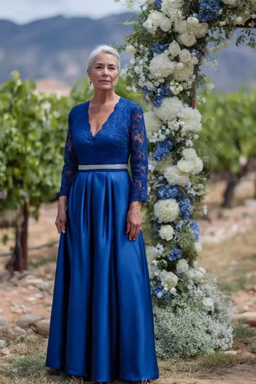 A woman over 50 wearing a royal blue A-line gown with lace sleeves and a silver belt, standing beside a floral arch in a vineyard setting, full body view 