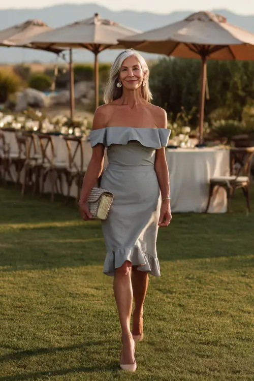 A woman over 50 wearing a soft grey off-the-shoulder cocktail dress with ruffled hem and clutch bag, walking across a lawn set up for an intimate summer wedding 