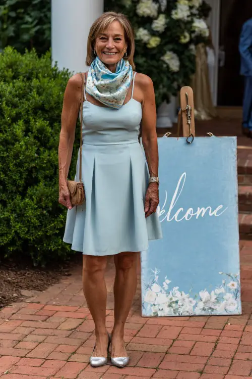 A woman over 60 in a light blue fit-and-flare dress with a floral scarf and silver flats, smiling near a decorative welcome sign at a summer wedding entrance 