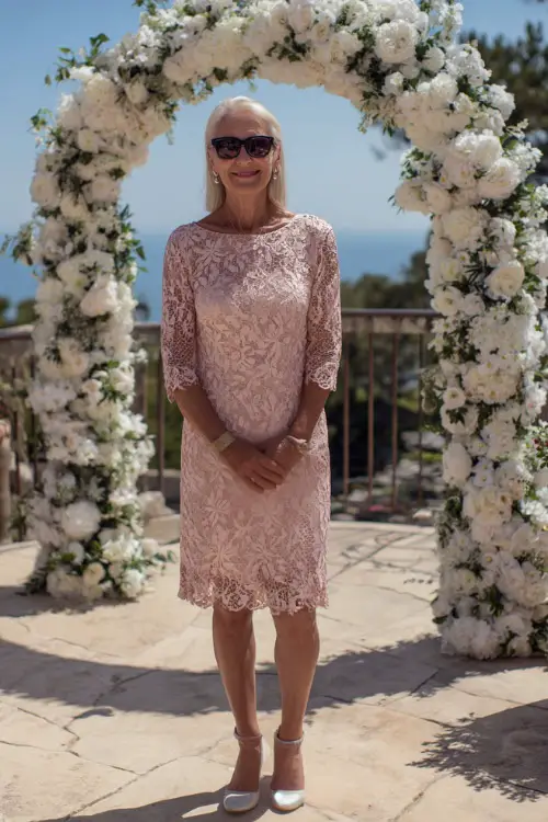 A woman over 60 in a soft blush pink A-line dress with lace overlay and block heels, posing beside a white floral arch at an outdoor ceremony, full body view 