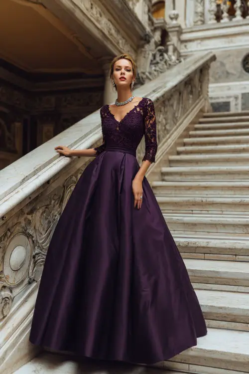 A woman wearing a deep purple floor-length gown with lace sleeves and silver jewelry, posing at the top of a grand staircase inside an elegant wedding venue 